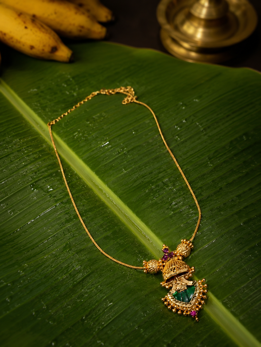 Traditional South Indian gold chain necklace featuring a Jhumka-style pendant with a vibrant green gemstone, displayed on a wet banana leaf next to yellow bananas.
