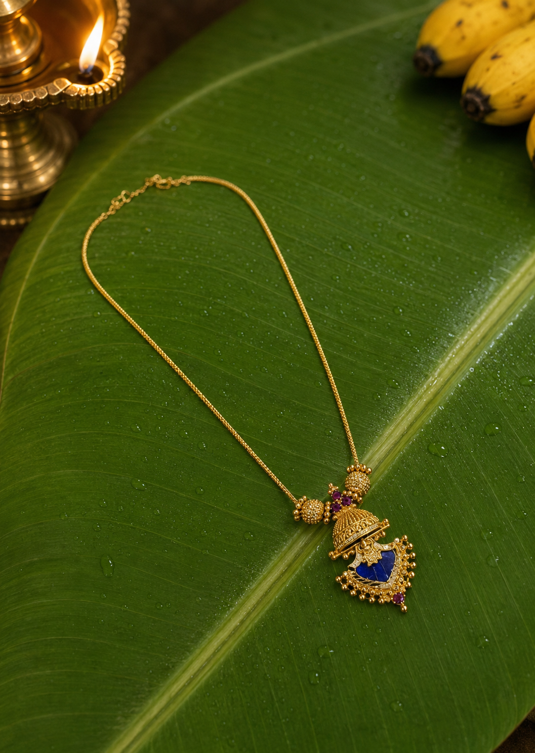 Kerala-style gold chain necklace with an intricate pendant inlayed with a deep blue sapphire-colored stone, arranged on a fresh green banana leaf with a lit brass diya lamp.