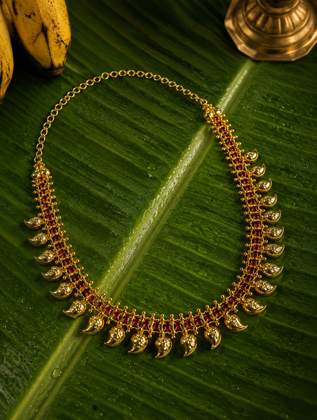 A traditional South Indian gold Manga Mala necklace studded with a row of vibrant red ruby gemstones and classic mango paisley motifs, beautifully arranged flat on a wet green banana leaf next to yellow bananas.
