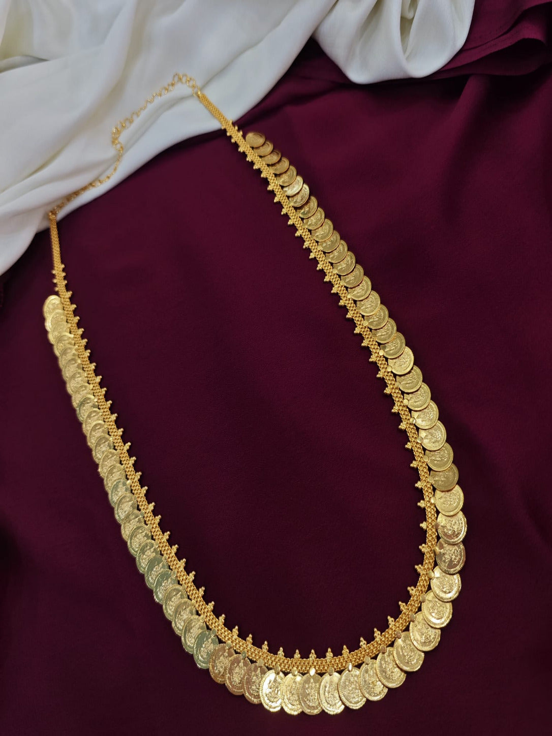 A long, traditional South Indian Kasu Mala gold necklace featuring a dense row of stamped Lakshmi coins attached to an intricate gold chain, elegantly draped over a deep maroon and off-white fabric background.
