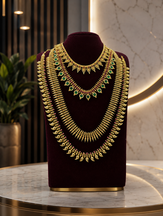 A premium multi-layered traditional South Indian bridal gold jewelry collection presented on a dark maroon bust display. The arrangement features an intricate leaf-motif gold choker, a necklace lined with vibrant green round stones, and three layered long traditional Kerala necklaces including spike (Mullamottu) and mango (Manga Mala) designs, set against an elegant interior background with warm ambient lighting.