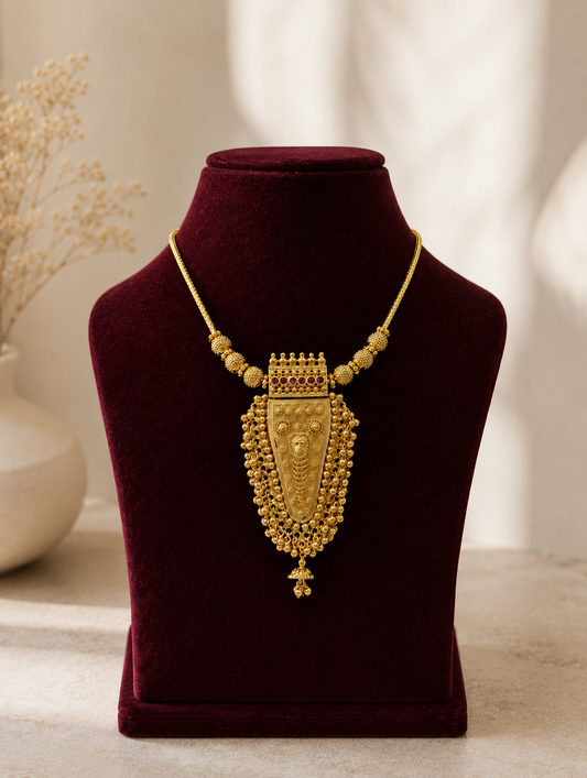 A close-up shot of an antique-finished gold necklace showcasing a large, detailed shield-shaped pendant with fine beadwork and a tiny jhumka droplet, beautifully presented on a dark maroon velvet jewelry bust under warm, natural lighting.