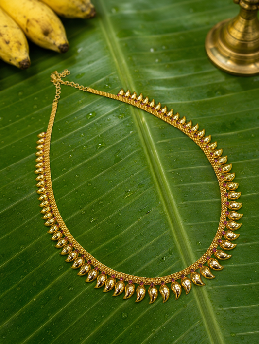 A traditional South Indian gold Manga Mala necklace adorned with a continuous row of polished mango-shaped paisley motifs and small crimson ruby-colored stones, laid flat across a wet green banana leaf featuring yellow bananas and a brass vilakku base in the upper corners.