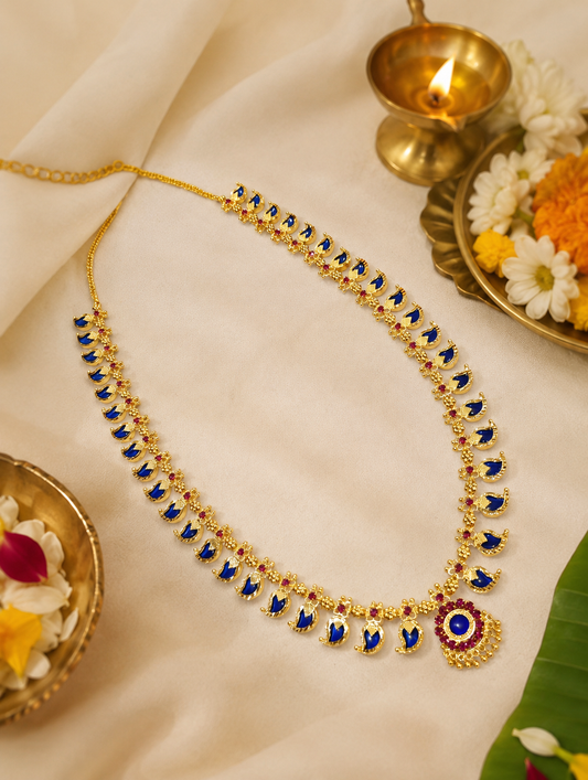 A traditional long Kerala Palakka Haram gold necklace inlaid with vibrant royal blue leaf-shaped stones and tiny pink ruby accents, elegantly placed on a soft off-white silk fabric alongside a lit brass diya and fresh marigold flowers.