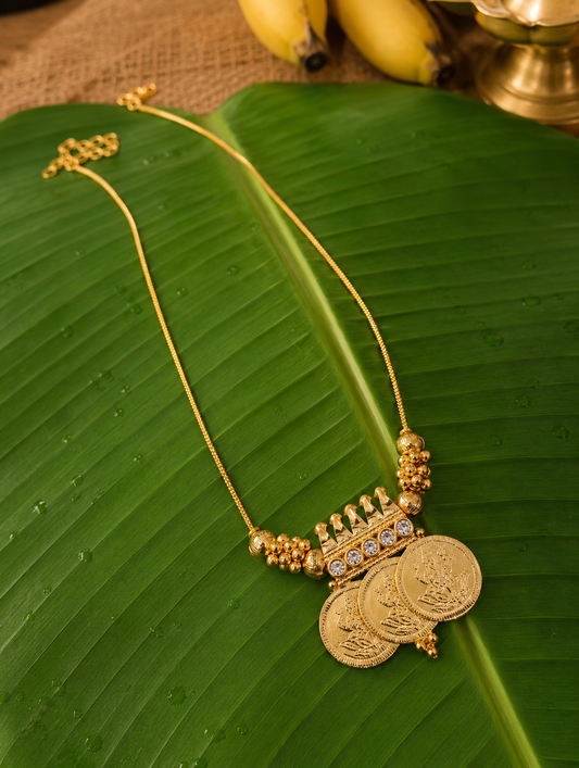 A traditional South Indian gold-toned chain necklace featuring a prominent pendant made of three overlapping coins embossed with Goddess Lakshmi, accented by a row of small clear white stones, displayed flat on a wet, vibrant green banana leaf.