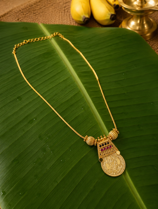 A traditional South Indian gold-toned chain necklace featuring an intricate trapezoidal pendant frame embedded with tiny colored stones, holding a round stamped Lakshmi coin, presented flat on a wet green banana leaf with yellow bananas and a brass diya in the background.