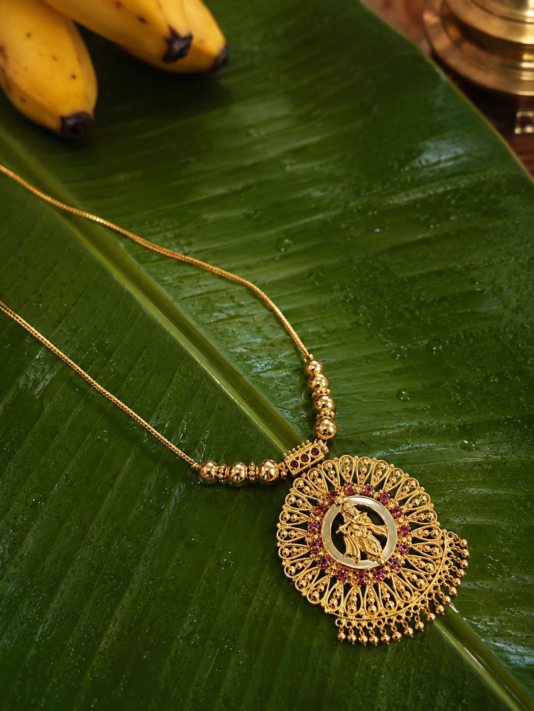 A traditional gold-toned Kanna necklace featuring a large circular filigree pendant with an engraving of Lord Krishna playing the flute, accented with small ruby stones and sleek gold beads, displayed elegantly on a wet green banana leaf with bananas in the background.