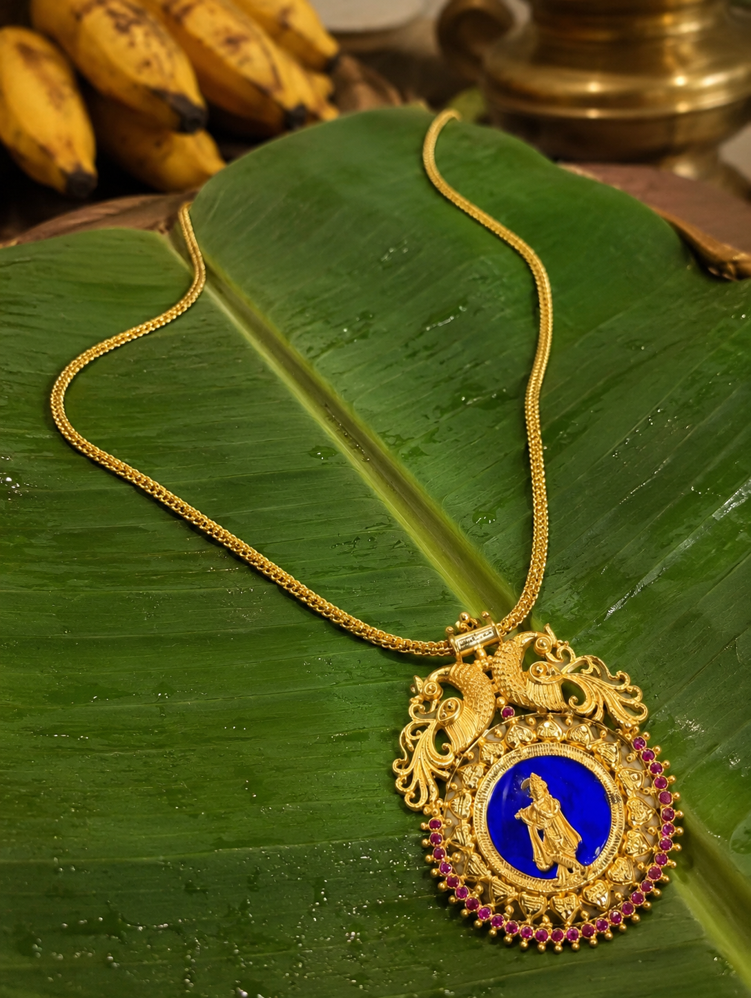 The traditional gold-toned Krishnaaa pendant necklace featuring an intricate engraving of Lord Krishna playing the flute over a vibrant blue enamel background, crowned with dual peacock motifs and framed with a row of small ruby stones on a fresh green banana leaf.