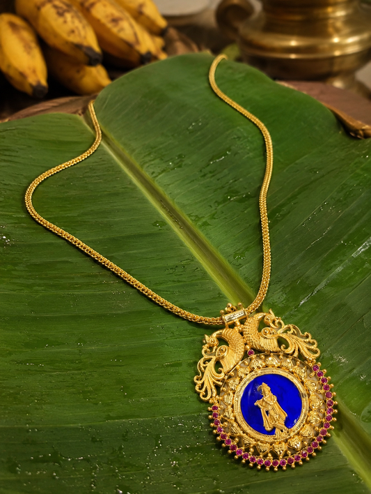 The traditional gold-toned Krishnaaa pendant necklace featuring an intricate engraving of Lord Krishna playing the flute over a vibrant blue enamel background, crowned with dual peacock motifs and framed with a row of small ruby stones on a fresh green banana leaf.