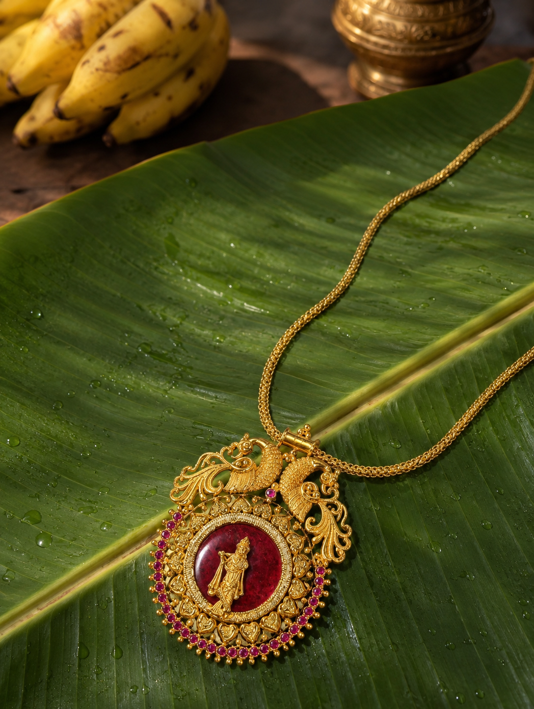 A variant of the Krishnaaa traditional gold necklace showcasing a deep crimson red enamel center behind the golden Lord Krishna figurine, detailed with twin golden peacocks and a pinkish-ruby stone border, resting on a banana leaf backdrop.