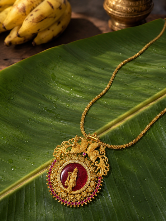 A variant of the Krishnaaa traditional gold necklace showcasing a deep crimson red enamel center behind the golden Lord Krishna figurine, detailed with twin golden peacocks and a pinkish-ruby stone border, resting on a banana leaf backdrop.