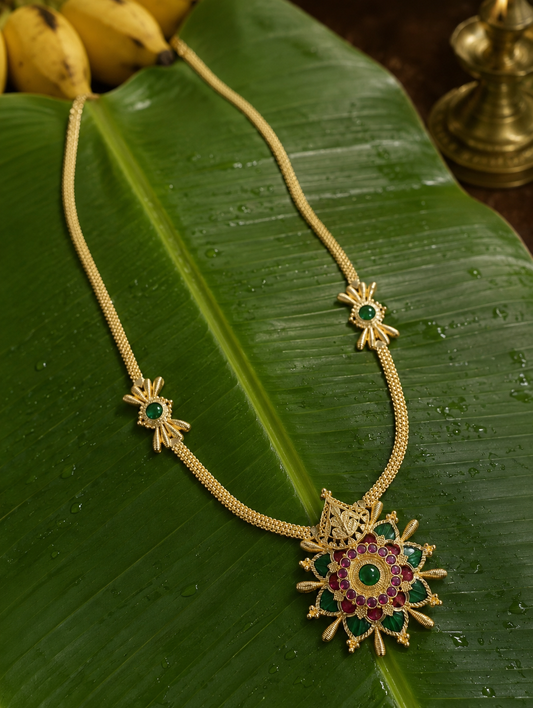 A traditional South Indian gold-toned necklace, featuring a prominent circular sunburst pendant set with ruby and emerald gemstones, accented by two emerald-centered side motifs along the intricate chain, displayed on a fresh green banana leaf.