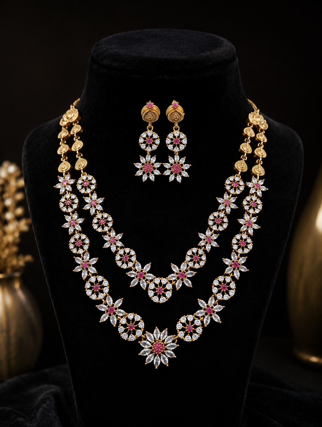 Elegant L005 bridal jewelry set showcasing a two-layered gold necklace and matching long drop earrings, intricately designed with white cubic zirconia floral patterns and pinkish-red ruby centers on a black velvet display bust.