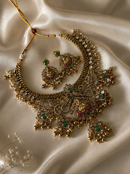 A traditional South Indian bridal jewelry set featuring a broad antique gold choker necklace and matching jhumka-style earrings with a central Goddess Lakshmi design, green and red stones, and hanging gold beads on a black display bust.