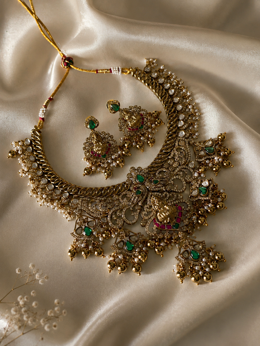A traditional South Indian bridal jewelry set featuring a broad antique gold choker necklace and matching jhumka-style earrings with a central Goddess Lakshmi design, green and red stones, and hanging gold beads on a black display bust.