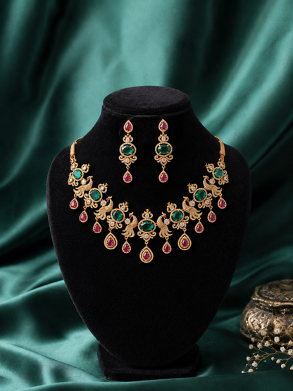 A traditional Victorian antique gold necklace and earring set featuring alternating peacock panels, large green emerald centers, and ruby teardrop pendants, arranged on emerald green satin fabric.