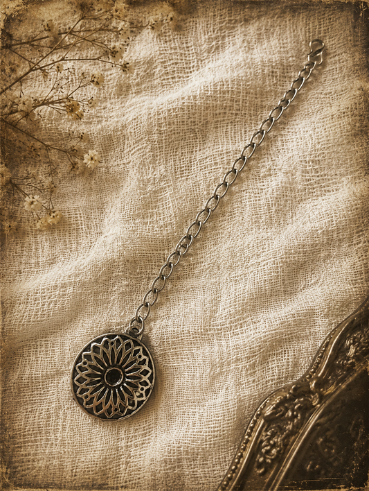 An overhead flat-lay shot of an oxidised silver maang tikka from the Gangu collection, featuring a circular, intricate openwork floral medallion attached to a metal link chain with a hook clasp. It is styled on a textured cream linen background with a vintage gold mirror edge.