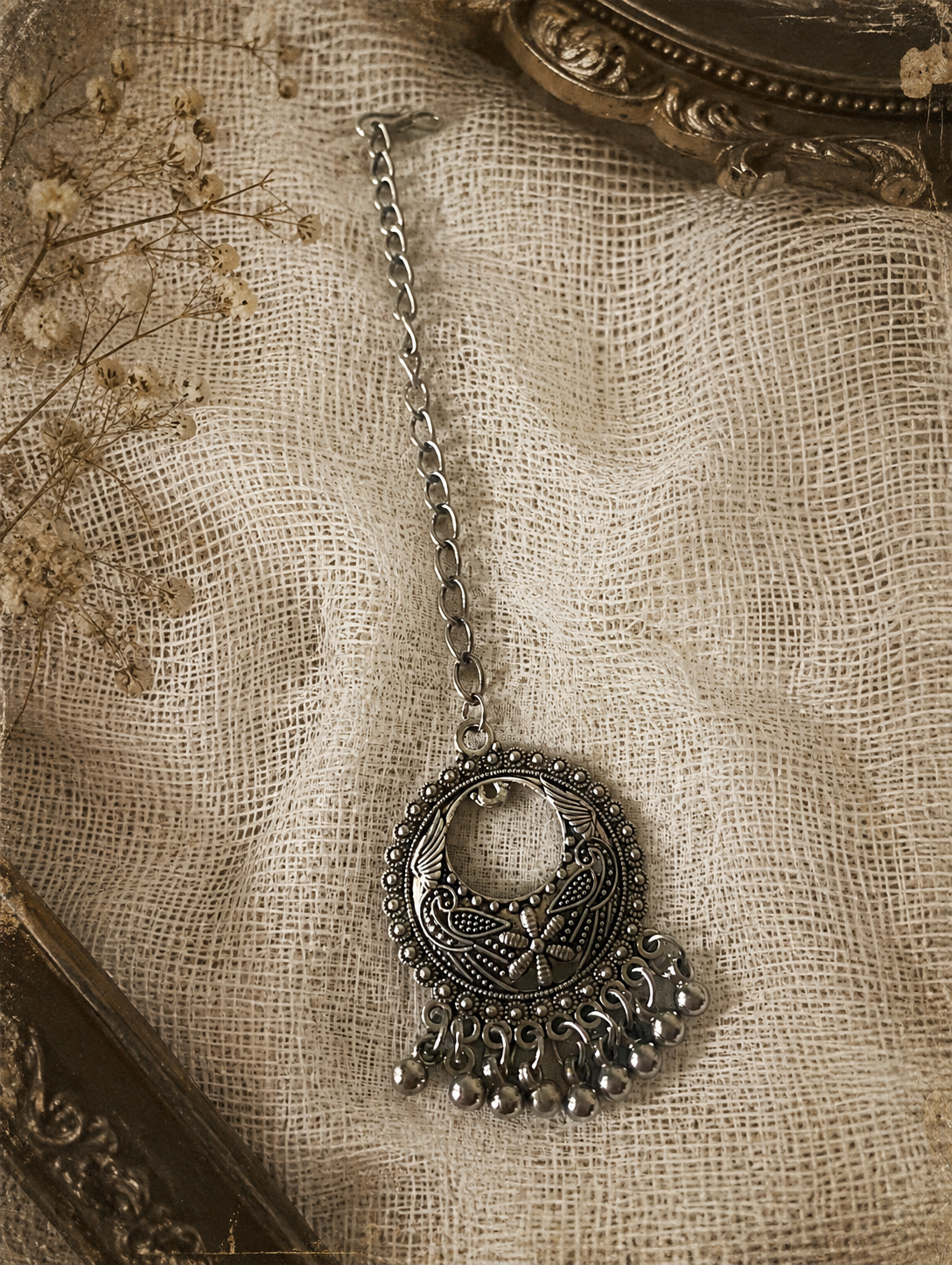 An overhead flat-lay shot of an oxidised silver maang tikka from the Magha collection. It features a crescent-shaped chandbali medallion engraved with fine peacock and floral patterns, finished with cascading silver ghungroo balls at the bottom, attached to a simple metal link chain on a textured cream mesh fabric backdrop.