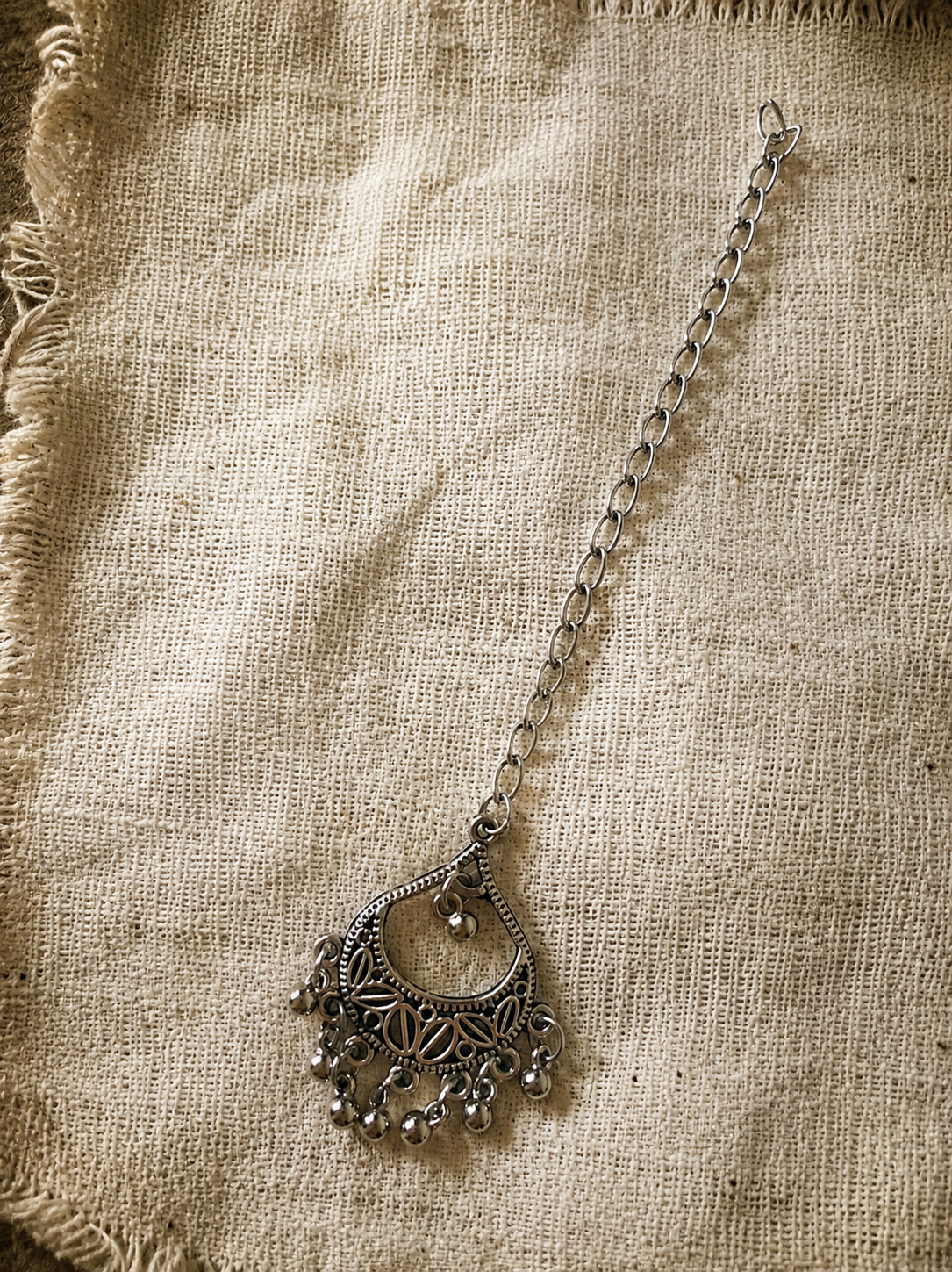 An overhead flat-lay shot of an oxidised silver maang tikka from the Parool collection. It features an openwork teardrop-shaped medallion detailed with small geometric leaf motifs and fringed with tiny metallic ghungroo balls, connected to a classic metal link chain on a coarse beige burlap background.