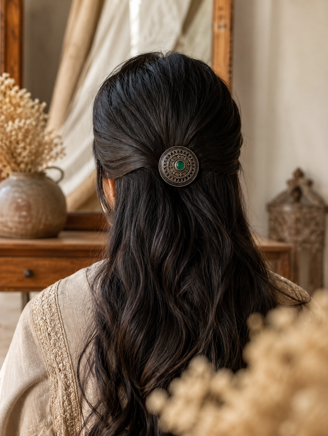 A back view of a woman with long, dark wavy hair styled in a elegant half-updo, secured with a circular oxidised silver hair pin from the Sunburst collection. The hair accessory features fine beaded detailing and a prominent round green stone at the center. The rustic background includes a wooden vanity and vintage mirror.