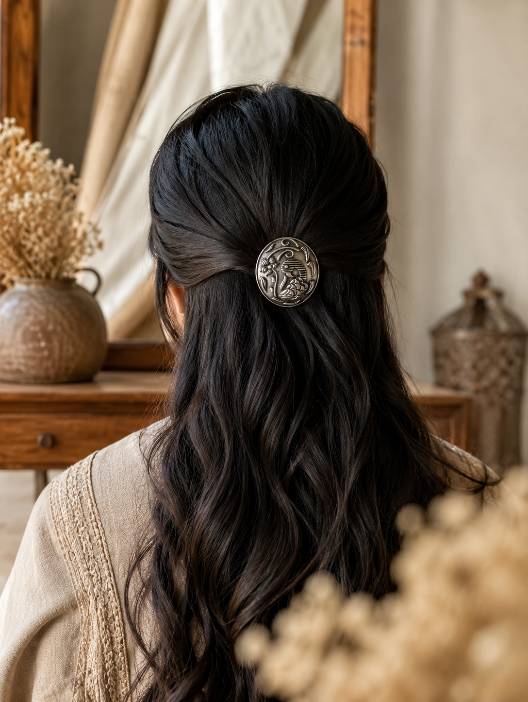 A back view of a woman with long, dark wavy hair styled in an elegant half-updo, fastened with an oval oxidised silver hair pin from the Peacock Punch collection. The accessory features a detailed embossed engraving of a traditional peacock and floral pattern, set against a rustic vanity background with a vintage mirror.