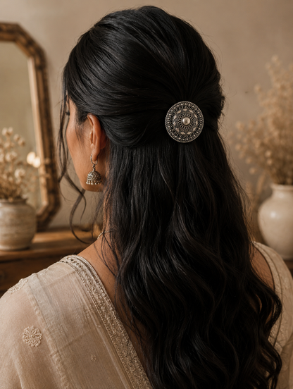 A back view of a woman with long, dark wavy hair styled in a graceful half-updo, secured with a minimalist hexagon-shaped silver hair pin. The accessory features a geometric hollow hexagonal outline detailed with delicate traditional metal beadwork along its edges.