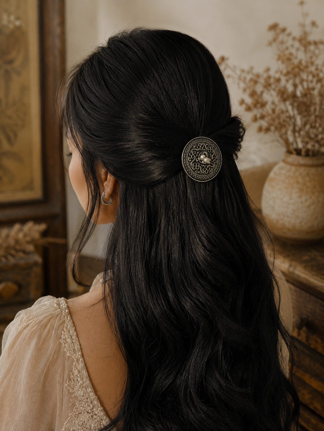 A back-angled view of a woman with long, dark wavy hair styled in a graceful half-updo, secured with a circular oxidised silver hair pin. The accessory features detailed ethnic filigree openwork work with small white pearls clustered at the center, set against a warm, rustic studio background.