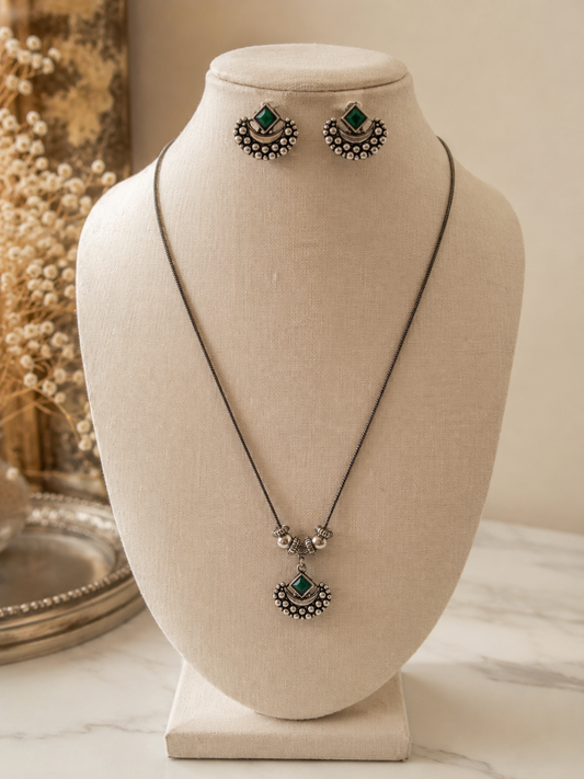 Oxidized silver jewelry set featuring a long chain pendant necklace and matching stud earrings, both adorned with a diamond-shaped green stone and a beaded crescent tribal design.