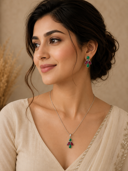 A woman modeling an oxidized silver jewelry set featuring a delicate chain necklace with a green and pinkish-ruby red leaf pendant, paired with matching drop earrings and a traditional cream saree.