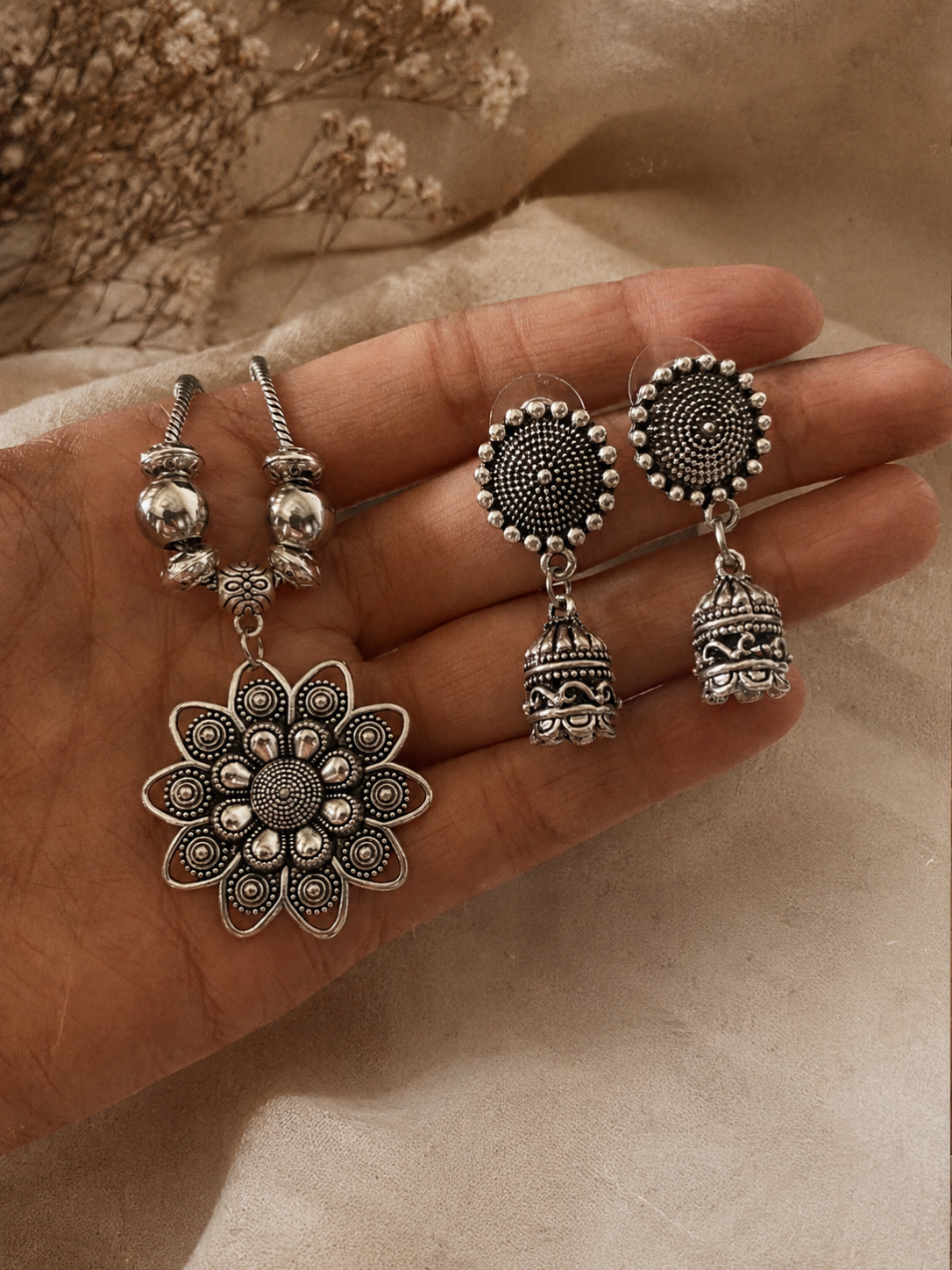 An oxidized silver jewelry set held in a hand, featuring a thick chain necklace with a large intricate floral mandala pendant and matching round stud jhumka dangle earrings.