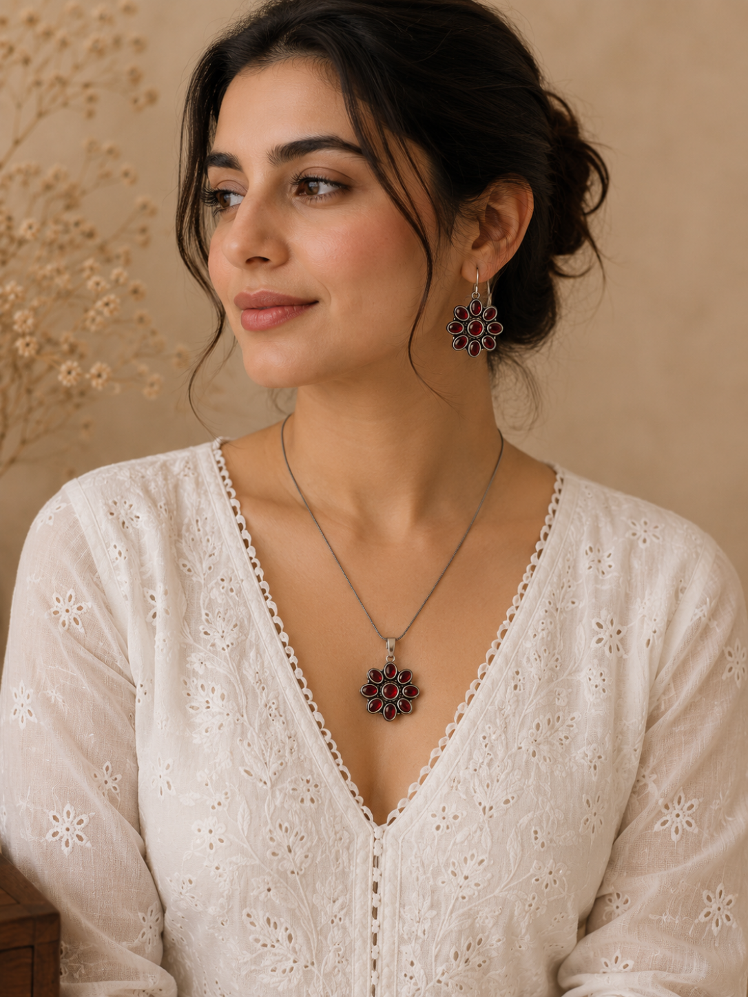 A woman modeling an oxidized silver jewelry set with a thin chain necklace and a matching floral-shaped pendant and hook earrings, all inset with deep ruby red glass stones, worn with a white embroidered chikankari top.