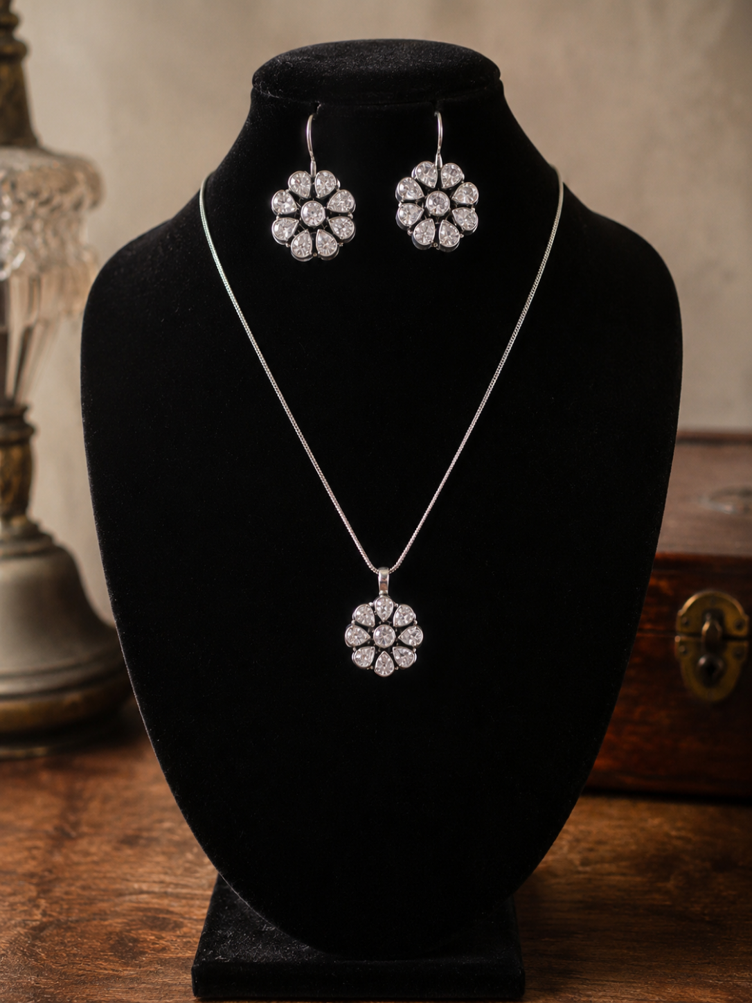 A sparkling oxidized silver jewelry set displayed on a black bust mannequin, featuring a delicate chain necklace with a floral-shaped clear stone pendant and matching dangle hook earrings.