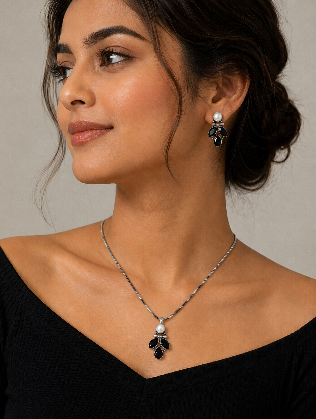 A woman modeling an oxidized silver jewelry set featuring a delicate chain necklace with a black stone leaf-motif pendant topped with a white pearl, paired with matching drop stud earrings and a black off-the-shoulder top.