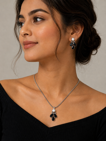 A woman modeling an oxidized silver jewelry set featuring a delicate chain necklace with a black stone leaf-motif pendant topped with a white pearl, paired with matching drop stud earrings and a black off-the-shoulder top.
