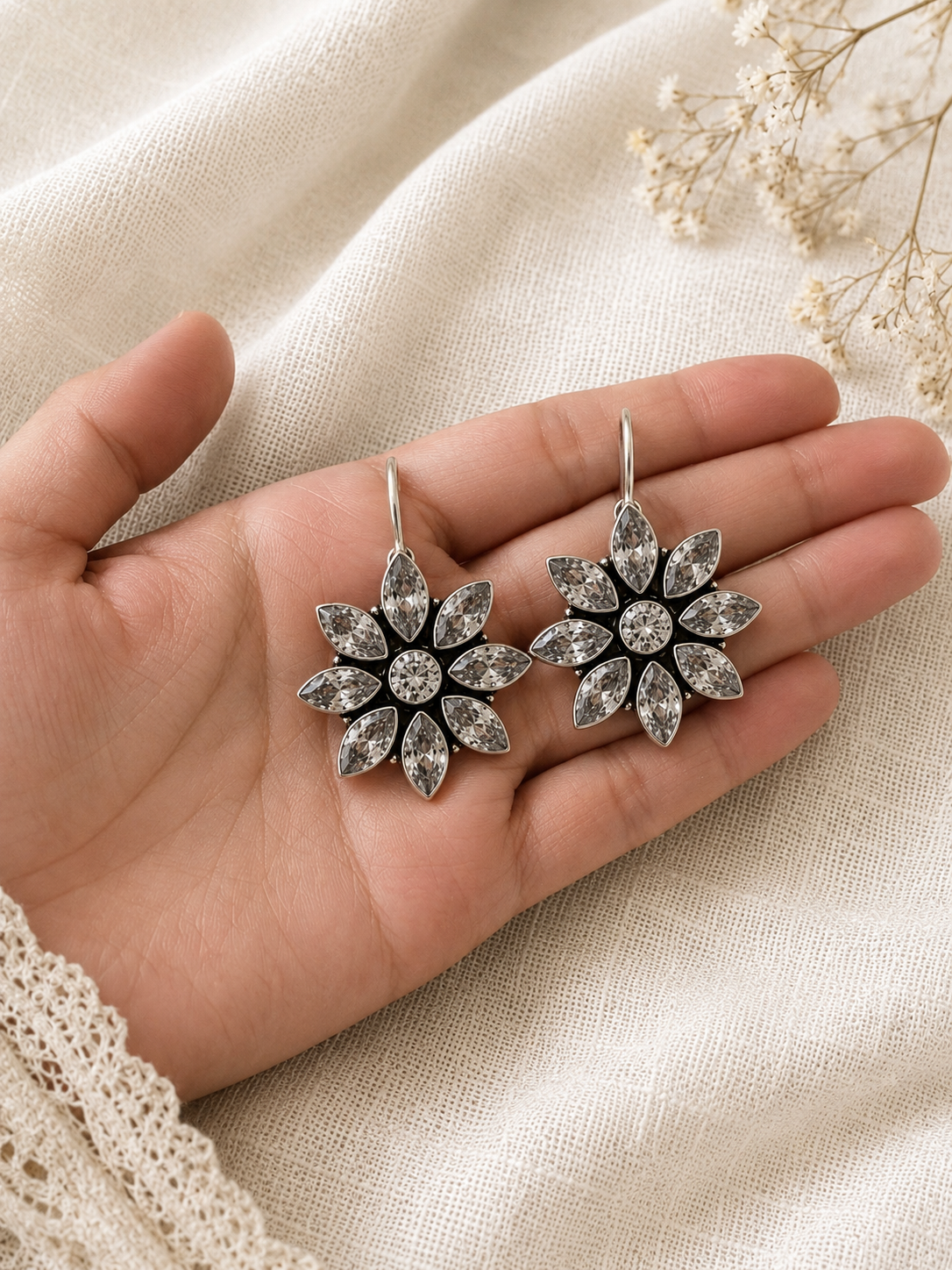 Flower-shaped silver earrings held in a hand on a beige fabric background
