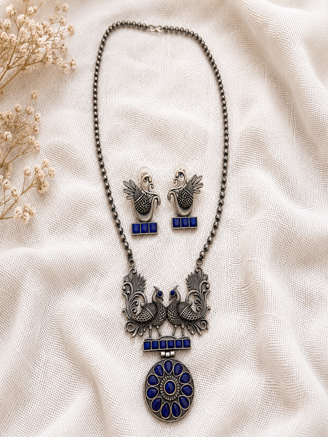 Oxidized silver long chain necklace featuring a twin peacock motif and a circular pendant inset with vibrant blue stones, alongside matching peacock stud earrings on a textured off-white fabric backdrop.