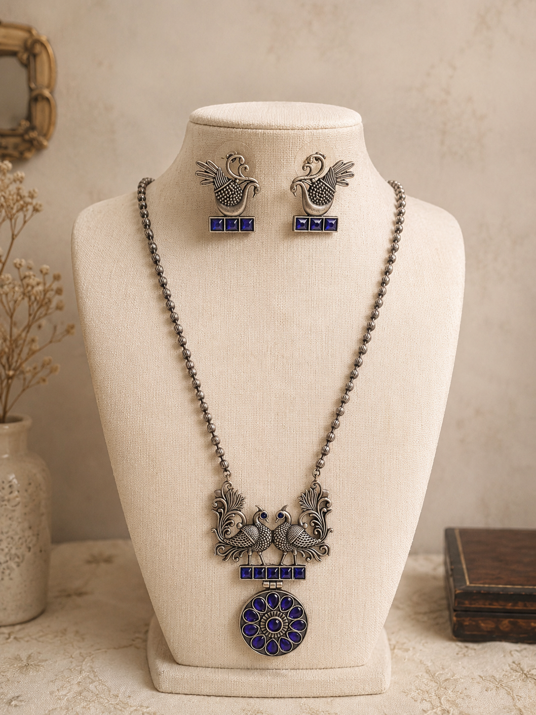 An oxidized silver long beaded chain necklace with a twin peacock and blue stone pendant, displayed on a cream-colored mannequin bust alongside matching blue stone peacock earrings.