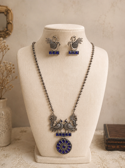 An oxidized silver long beaded chain necklace with a twin peacock and blue stone pendant, displayed on a cream-colored mannequin bust alongside matching blue stone peacock earrings.