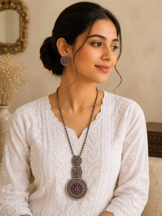 A smiling woman wearing a white embroidered chikankari kurta, styled with a long oxidized silver beaded chain necklace featuring a three-tier circular mandala pendant inset with pink stones, and matching round stud earrings.