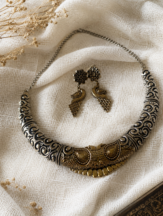 Premium flat-lay of a dual-tone oxidized silver and gold antique Hasli choker necklace paired with matching peacock dangle earrings on a textured white linen fabric.