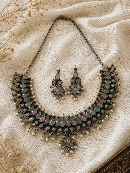 An oxidized silver collar necklace and matching drop earrings set with pastel pink and mint green teardrop stones and tiny pearl accents, laid flat on a textured cream linen background.