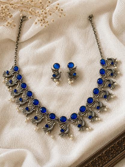 An oxidized silver collar necklace featuring a row of vibrant round royal blue stones with hanging peacock motifs and pearl droplets, shown with matching blue stone peacock earrings on a textured cream fabric backdrop.