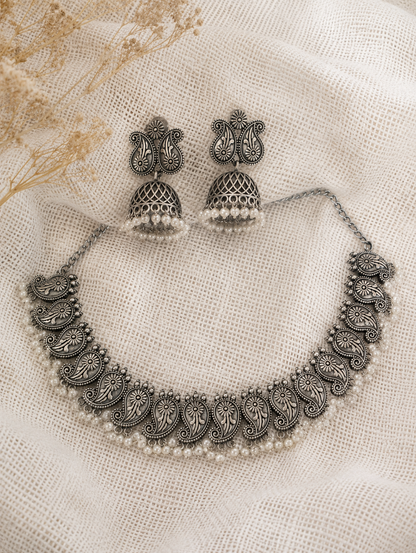 An oxidized silver choker necklace featuring a repeating paisley motif and a delicate lower border of small white pearls, paired with matching paisley stud jhumka earrings on a textured cream mesh fabric backdrop.