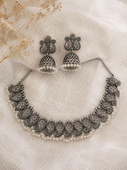 An oxidized silver choker necklace featuring a repeating paisley motif and a delicate lower border of small white pearls, paired with matching paisley stud jhumka earrings on a textured cream mesh fabric backdrop.
