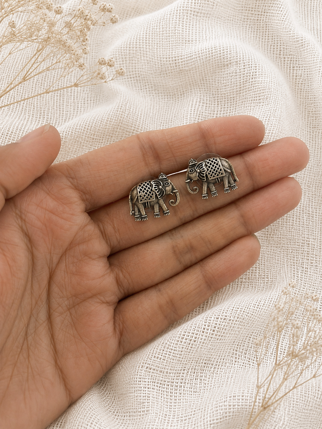 Two silver elephant earrings held between fingers on a light fabric background