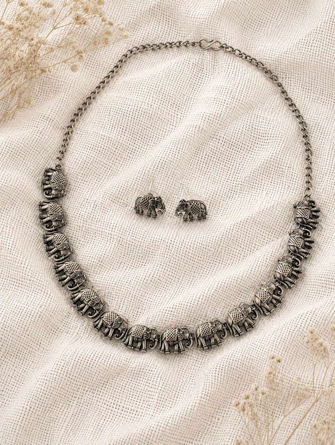 An oxidized silver collar necklace composed of a linked row of finely detailed antique elephant charms, displayed with matching elephant stud earrings on a textured off-white mesh fabric background.