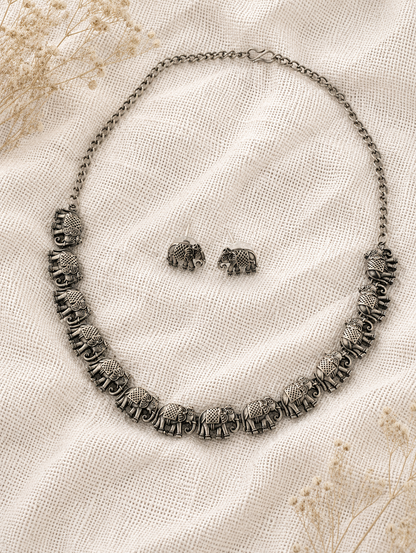 An oxidized silver collar necklace composed of a linked row of finely detailed antique elephant charms, displayed with matching elephant stud earrings on a textured off-white mesh fabric background.