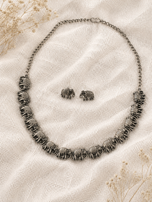 An oxidized silver collar necklace composed of a linked row of finely detailed antique elephant charms, displayed with matching elephant stud earrings on a textured off-white mesh fabric background.
