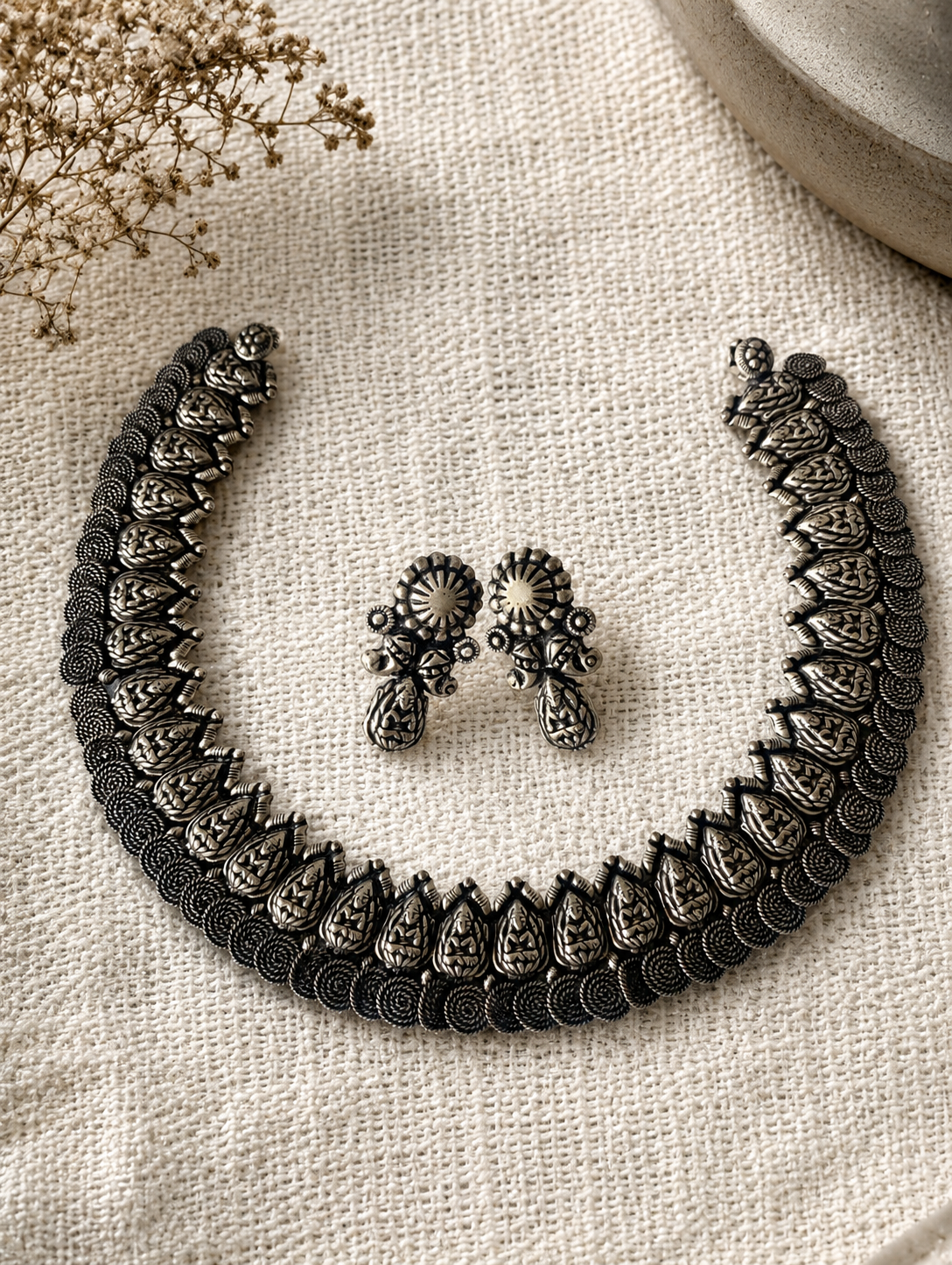 An antique oxidized silver jewelry set featuring a broad, rigid choker necklace and matching stud earrings carved with traditional Indian goddess motifs, arranged on a textured off-white fabric backdrop.