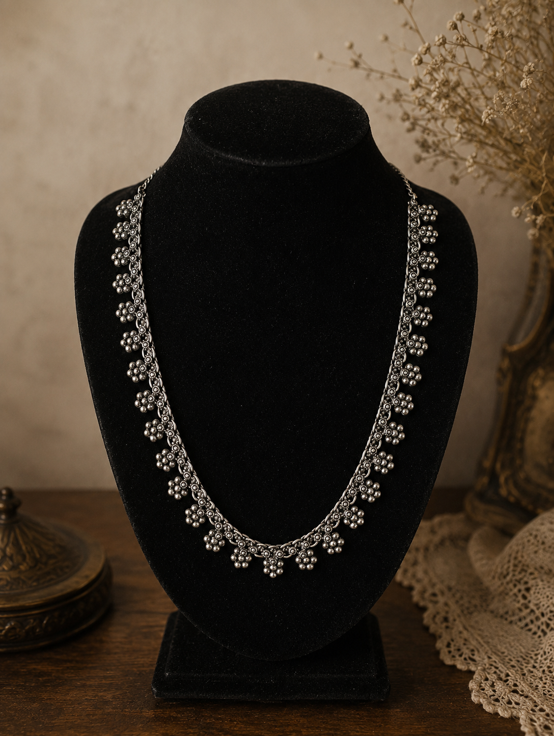 An antique-style oxidized silver necklace featuring intricate, repeating floral cluster charms, showcased on a black velvet bust display against a rustic backdrop.